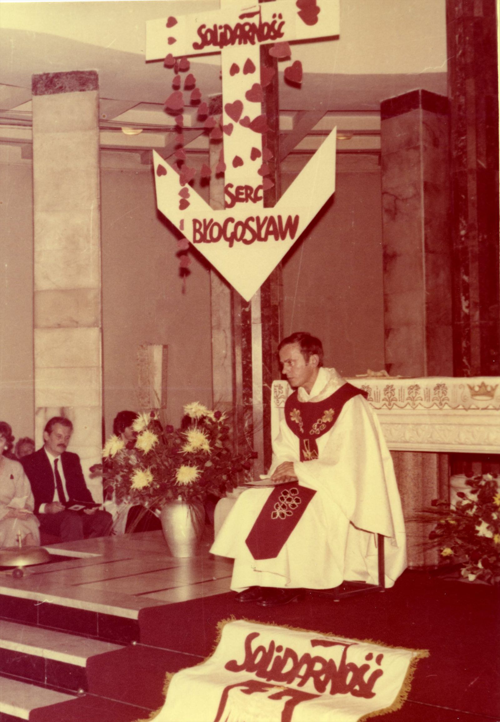 Служба впомещении металлургического завода «Хута Варшава» в первую годовщину августовской забастовки в Гданьске. Отец Ежи облачен в праздничную литургическую одежду (казулу), над импровизированным алтарем висит инсталляция с логотипом «Солидарности» и надписью «Благослови сердце». 31 августа 1981 года. Варшава. Фото из архивов института национальной памяти, Польша.