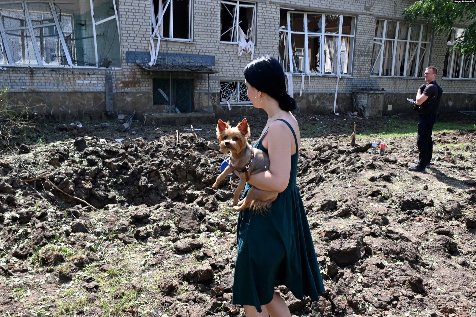 Женщина с собакой стоит у огромной воронки, образовавшейся в результате ракетного удара рядом с разрушенным зданием школы в Харькове. 4 июля / Sergey Bobok / AFP / Radio Svoboda​