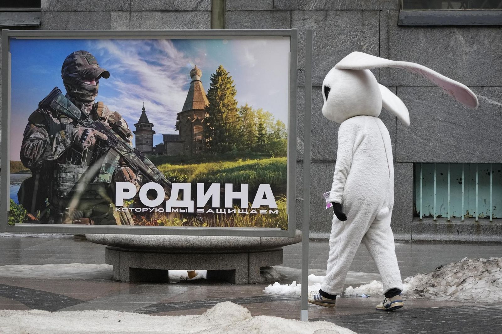 Промоутер, приглашающий прохожих посетить музей, проходит мимо баннера с изображением российского военного и надписью «Родина, которую мы защищаем» на уличной выставке военных фотографий в Санкт-Петербурге. 14 марта / Dmitri Lovetsky / AP Photo 